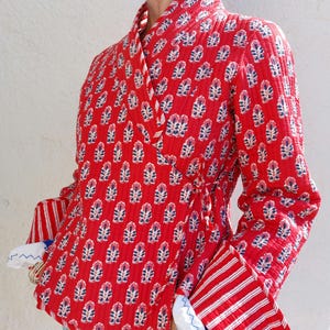 Könnte beinhalten: Rote, gesteppte Wickeljacke mit einem Blumenmuster in Weiß und Blau. Die Jacke hat lange Ärmel mit rot-weiß gestreiften Manschetten und einen Bindverschluss in der Taille. Wickeljacke.