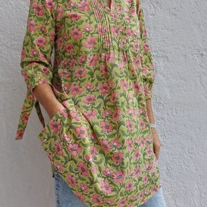 May include: A tunic top featuring a light green base with a repeating pink floral pattern, a split V-neckline with vertical pleats, three-quarter length sleeves with tie-cuff details, and two large patch pockets on the front of the garment.