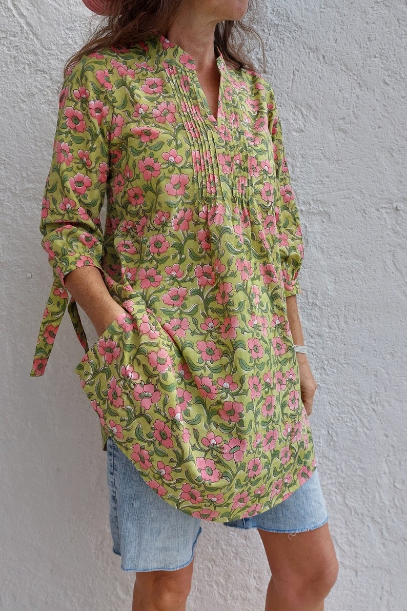 May include: A tunic top featuring a light green base with a repeating pink floral pattern, a split V-neckline with vertical pleats, three-quarter length sleeves with tie-cuff details, and two large patch pockets on the front of the garment.