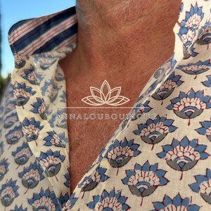 May include: A button-down shirt featuring a repeating blue and grey floral pattern on an off-white background. The inner collar displays a contrasting blue, white, and pink striped fabric. The text ANNALOUBOUTIQUE is printed in white.