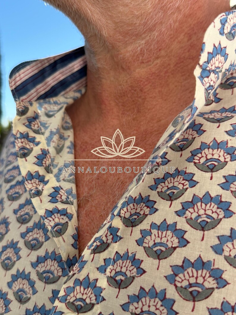 May include: A button-down shirt featuring a repeating blue and grey floral pattern on an off-white background. The inner collar displays a contrasting blue, white, and pink striped fabric. The text ANNALOUBOUTIQUE is printed in white.