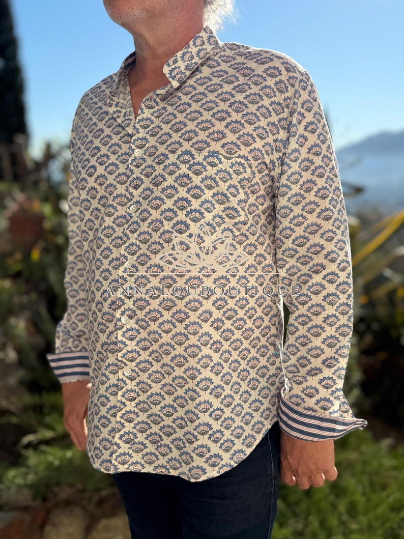 May include: A long-sleeved button-down shirt featuring a repeating blue and muted red floral block print on a cream background. The shirt includes a chest pocket, a standard collar, and cuffs with a blue and white striped pattern on the interior.