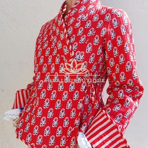 Peut inclure: Une veste portefeuille matelassée rouge vif ornée d'un motif floral bleu et blanc répété. La veste présente des manches longues avec de larges poignets rayés rouges et blancs qui se replient pour révéler une doublure blanche avec des coutures bleues.