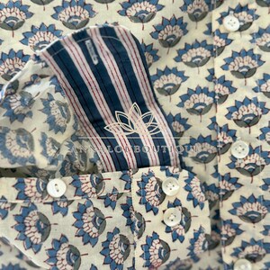 May include: A long-sleeve shirt featuring an off-white base with a repeating blue and grey floral pattern. The cuff interior displays blue and white vertical stripes with thin red lines. The shirt includes white buttons and the text ANNALOUBOUTIQUE.