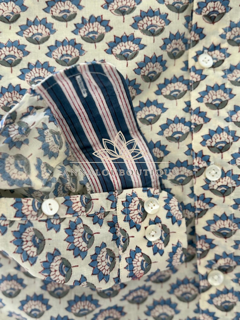 May include: A long-sleeve shirt featuring an off-white base with a repeating blue and grey floral pattern. The cuff interior displays blue and white vertical stripes with thin red lines. The shirt includes white buttons and the text ANNALOUBOUTIQUE.