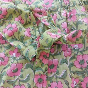 May include: A light green long-sleeved shirt featuring a repeating floral pattern of pink flowers with dark green leaves and stems. The garment includes a buttoned cuff on the sleeve, a fabric tie detail, and vertical pleats along the upper chest area.