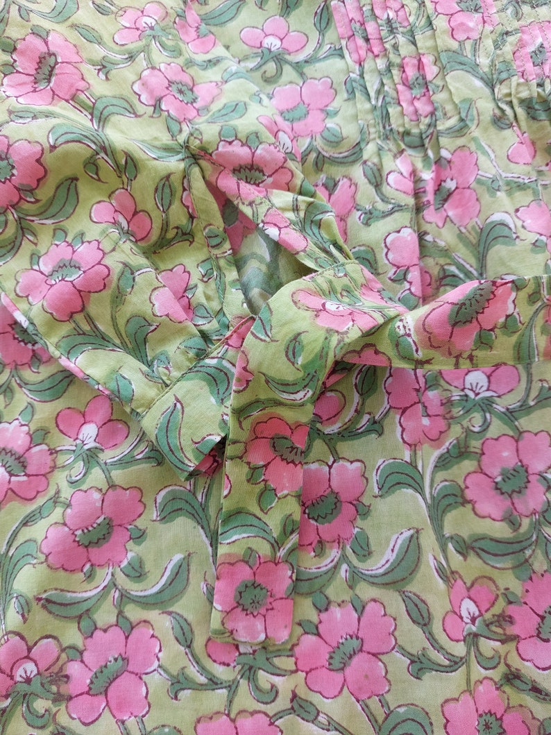 May include: A light green long-sleeved shirt featuring a repeating floral pattern of pink flowers with dark green leaves and stems. The garment includes a buttoned cuff on the sleeve, a fabric tie detail, and vertical pleats along the upper chest area.