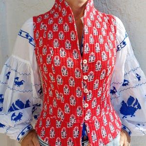 Peut inclure: Un gilet matelassé rouge avec un motif floral blanc et bleu. Le gilet a un col montant et est boutonné sur le devant. La blouse blanche en dessous a des manches avec des motifs d'oiseaux bleus.