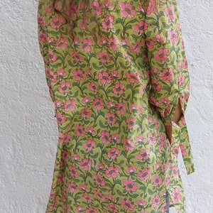 May include: A long-sleeved tunic featuring a green background with a repeating pink floral pattern, paired with blue denim shorts and a pink wide-brimmed hat. The tunic has side slits and tie details on the sleeves, providing a loose fit.