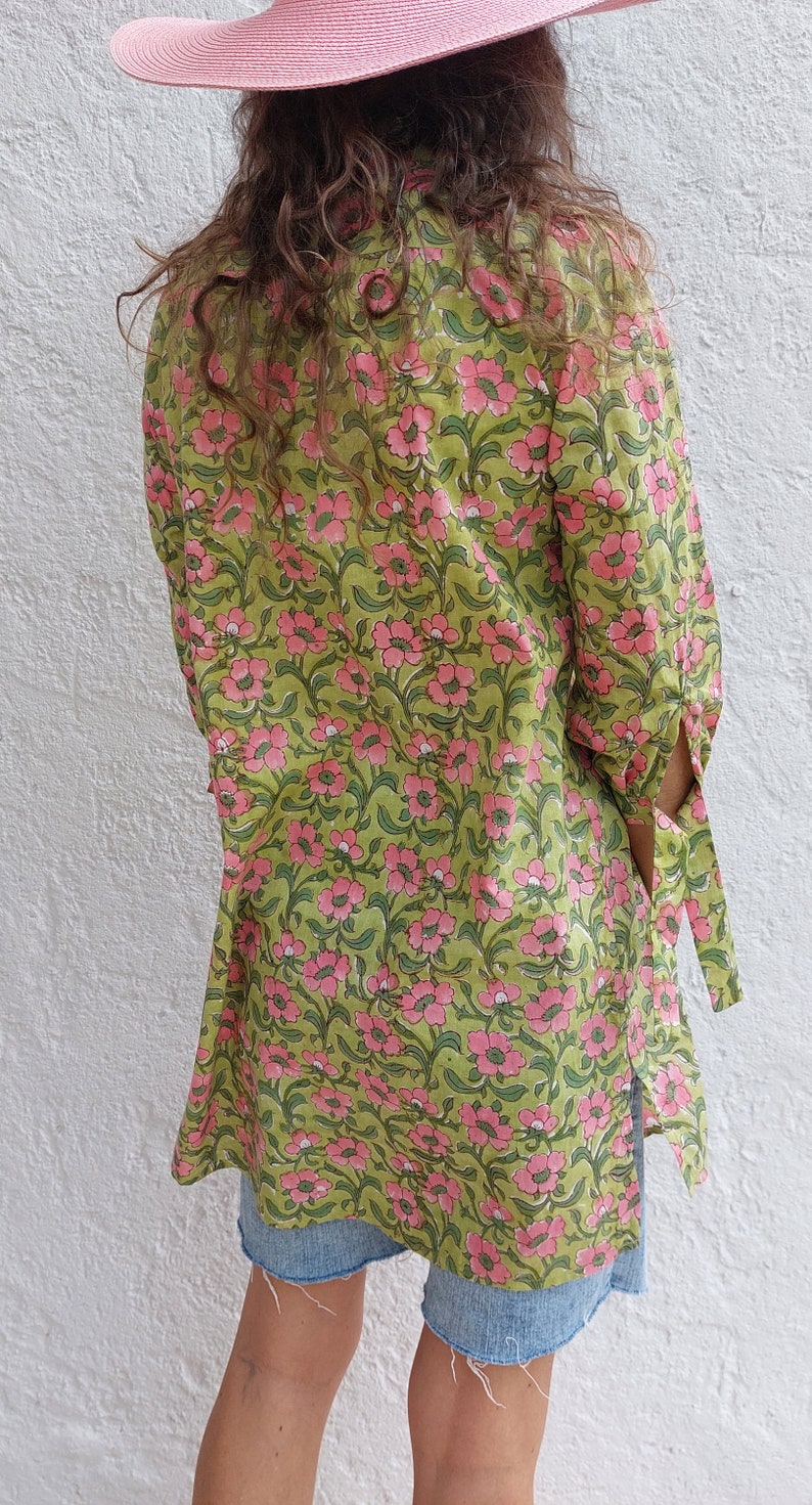 May include: A long-sleeved tunic featuring a green background with a repeating pink floral pattern, paired with blue denim shorts and a pink wide-brimmed hat. The tunic has side slits and tie details on the sleeves, providing a loose fit.