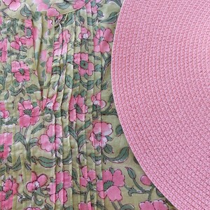 May include: A light green tunic featuring a pink floral pattern with vertical pleats along the front neckline and a round, textured pink woven placemat positioned to the right side of the garment, partially overlapping the floral fabric design.