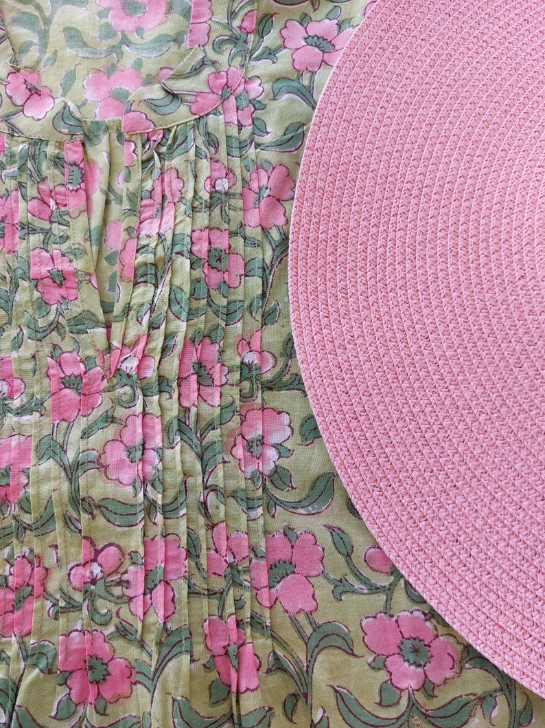 May include: A light green tunic featuring a pink floral pattern with vertical pleats along the front neckline and a round, textured pink woven placemat positioned to the right side of the garment, partially overlapping the floral fabric design.