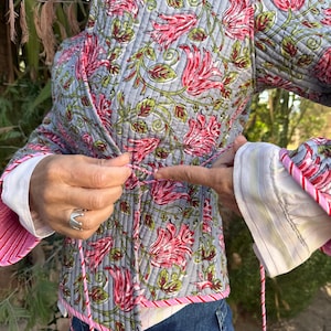 Peut inclure: Veste matelassée à motifs floraux roses, rouges et verts sur fond gris. La veste a une bordure rose et une fermeture à nouer. Les manches ont un poignet rayé blanc et rose.