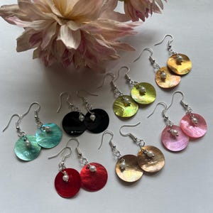 May include: A selection of handmade earrings in diverse colours, such as black, red, pink, and green. Each earring has a round, iridescent shell disc with a small, white pearl and silver-tone hooks. A pink flower is in the background.