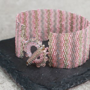 May include: A pink and silver beaded bracelet with a decorative clasp. The bracelet is made with small, seed beads and has a striped pattern.