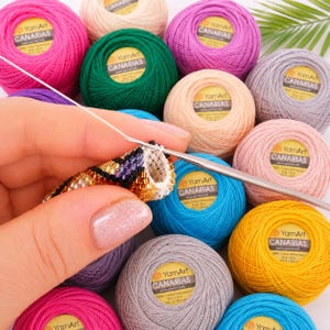 Peut inclure: Un crochet argenté travaille sur un projet de tube en perles noires, dorées et blanches. Le projet est entouré de plusieurs pelotes de fil YamArt Canarias 100% coton mercerisé dans diverses couleurs dont rose, vert, violet et jaune.