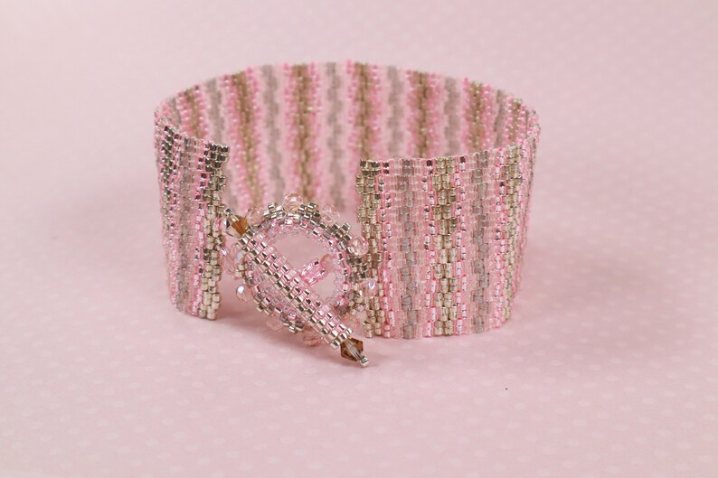 May include: A pink and silver beaded bracelet with a decorative clasp. The bracelet is made of small, square beads that are arranged in a striped pattern.