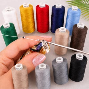 May include: A collection of twelve spools of thread in various colors including white, yellow, red, dark blue, light blue, green, brown, gray, and black, each labeled 100% PES or jeansová nit, alongside a metal crochet hook working on beadwork.