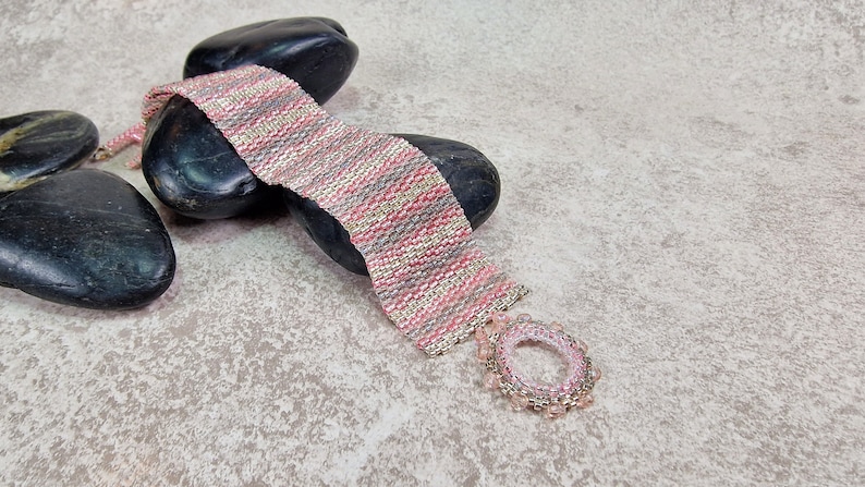 May include: A pink, gray, and silver beaded bracelet with a circular pendant. The bracelet is lying on top of smooth, black stones.