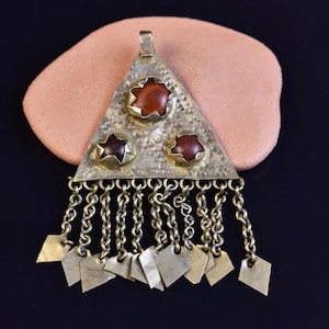 Puede incluir: Colgante de estilo vintage con base triangular de latón, adornado con tres piedras preciosas de color marrón rojizo. El colgante presenta cadenas colgantes con pequeños dijes de latón en forma de diamante. La pieza está sobre un fondo color melocotón.