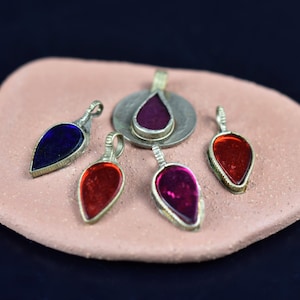 Puede incluir: Una colección de colgantes en forma de lágrima con engastes de metal plateado. Los colgantes presentan piedras translúcidas en tonos azul, rojo y morado. Se exhiben sobre una superficie rosa texturizada.