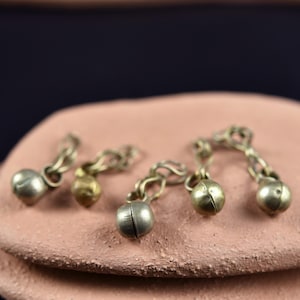 Può includere: Primo piano di diverse piccole campane in metallo color argento a forma di sfera, collegate da delicate catene. Le campane poggiano su una superficie strutturata marrone rossiccio chiaro, con uno sfondo blu scuro. Le campane hanno un aspetto vintage o antico.