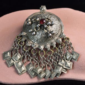 Può includere: Ciondolo in argento ornato con una gemma rossa centrale. Il ciondolo presenta intricati dettagli in rilievo e ciondoli quadrati pendenti. Accenti dorati e catenelle aggiungono un tocco vintage. Il ciondolo è esposto su una superficie rosa.