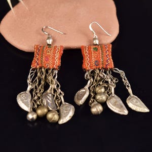 Puede incluir: Un par de pendientes hechos a mano con cuadrados de tela naranja con bordados coloridos. Cadenas plateadas cuelgan de los cuadrados, adornadas con dijes plateados en forma de hoja y pequeñas cuentas redondas. Cierre de gancho.