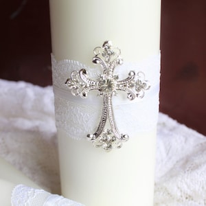 May include: Two white wedding candles with lace and a silver cross with rhinestones. The candles are on a white lace tablecloth.