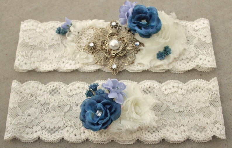 Puede incluir: Dos ligas de encaje blanco con adornos de flores azules y blancas. La liga superior tiene una pieza decorativa dorada y de perlas en el centro.