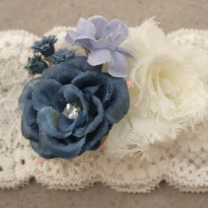 Puede incluir: Una liga de encaje blanco con un adorno de flores azules y blancas. La flor tiene un centro de pedrer&iacute;a.