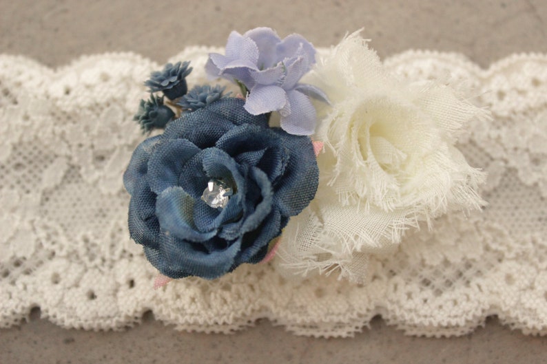 Puede incluir: Una liga de encaje blanco con un adorno de flores azules y blancas. La flor tiene un centro de pedrer&iacute;a.