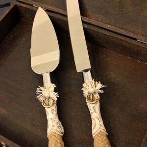 May include: A set of two cake knives with decorative handles. The handles are wrapped in burlap and lace with a pearl and anchor charm. The knives are in a wooden box.