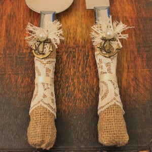 May include: A pair of cake cutting knives with burlap and lace handles. Each handle has a small anchor charm and a pearl accent.