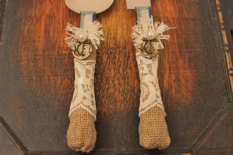 May include: A pair of cake cutting knives with burlap and lace handles. Each handle has a small anchor charm and a pearl accent.