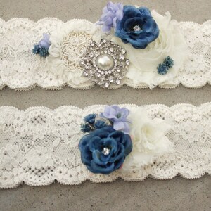 Puede incluir: Dos ligas de encaje blanco con flores de tela azul y blanco y un adorno de pedrer&iacute;a.