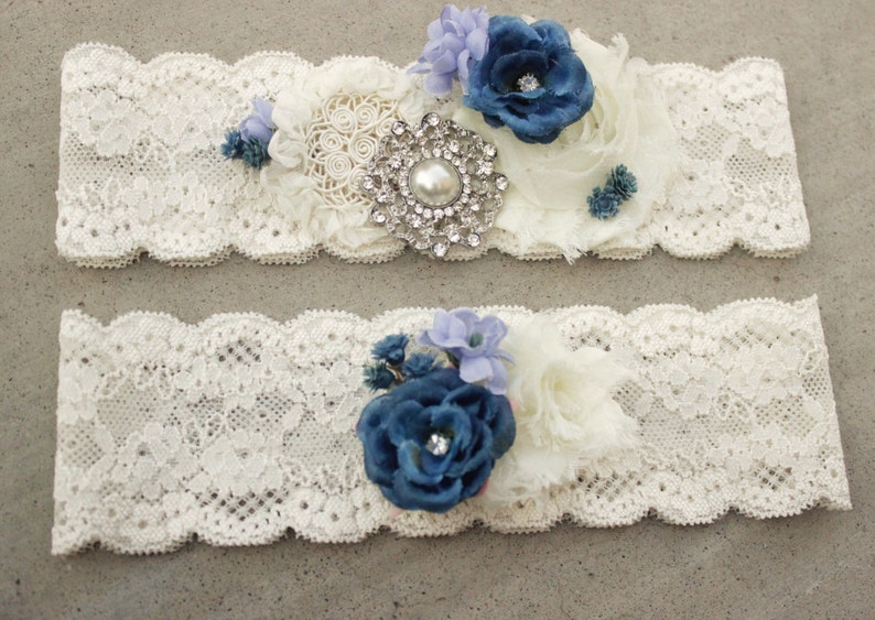 Puede incluir: Dos ligas de encaje blanco con flores de tela azul y blanco y un adorno de pedrer&iacute;a.