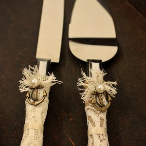 May include: A pair of wedding cake knives with white lace and burlap handles. Each handle features a small anchor charm with a pearl accent.