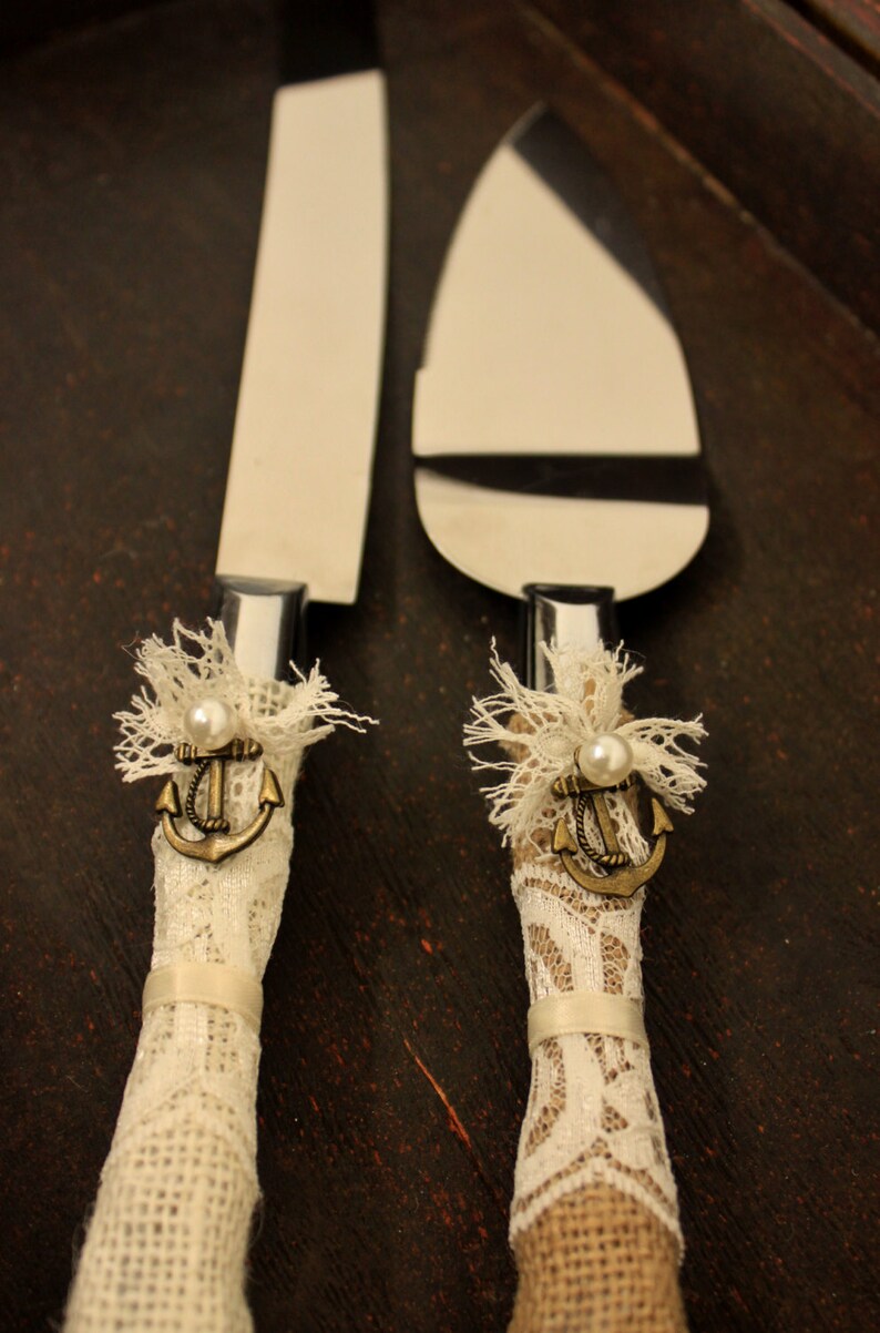 May include: A pair of wedding cake knives with white lace and burlap handles. Each handle features a small anchor charm with a pearl accent.