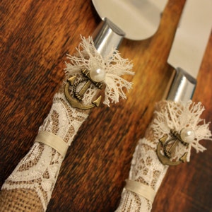 May include: A set of two cake knives with burlap and lace handles. Each handle features a small anchor charm and a pearl accent. The knives are perfect for cutting a wedding cake.