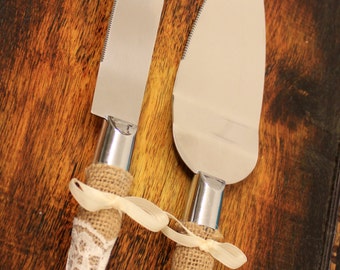May include: Two silver cake knives with burlap and lace handles. The knives are on a dark wood surface.