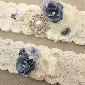 Puede incluir: Dos ligas de encaje blanco con flores de tela azul y blanco y un adorno de pedrer&iacute;a.