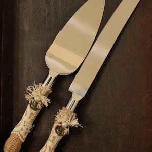 May include: A set of two silver cake knives with decorative burlap and lace handles. The handles feature a pearl and an anchor design.
