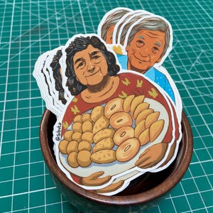 May include: A pile of stickers depicting a woman holding a plate of food. The woman is wearing a red patterned shirt. The stickers are in a wooden bowl, on a green grid surface. The stickers have a white border.