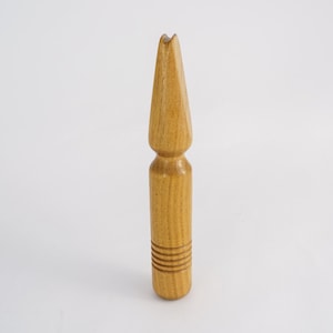 May include: A light brown wooden tool with a pointed top and a cylindrical body. The tool has a series of horizontal grooves near the base. The top has a split design. The tool is approximately 6 inches tall.