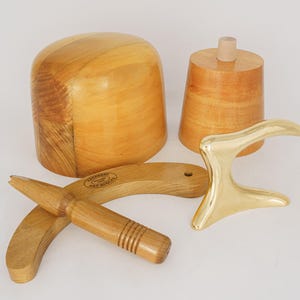 Könnte beinhalten: Eine Sammlung von Holzwerkzeugen zur Hutmacherei, darunter ein Hutblock, eine konische Form, ein kleines zylindrisches Stück, ein gebogenes Holzwerkzeug und ein goldfarbenes Metallwerkzeug. Die Werkzeuge sind auf einer weißen Oberfläche angeordnet.