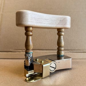 May include: A wooden and metal book press with a light-colored wooden handle and two turned wooden legs. The base is rectangular with brass and silver-colored metal components. The press is designed for applying pressure.