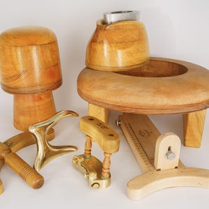 Könnte beinhalten: Eine Sammlung von Holzwerkzeugen zur Hutmacherei, darunter ein Hutblock, ein Hutspanner und verschiedene Formwerkzeuge. Die Werkzeuge sind aus hellem Holz und Messing gefertigt und haben eine natürliche Oberfläche. Der Hutblock ist etwa 20 cm hoch.