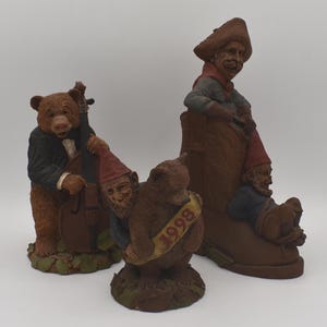 May include: Three whimsical figurines: a bear playing a cello, a bear hugging a gnome with a banner reading "1998", and a man in a cowboy hat seated in a boot with two gnomes. The figurines are brown and have detailed features.