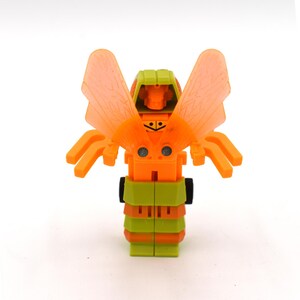 May include: A small, orange and green toy robot with translucent orange wings. The toy has a blocky design with visible joints and details, and is set against a white background. The toy's design is reminiscent of a transforming robot.